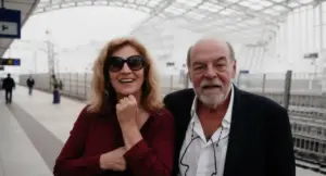 Due persone sorridenti in posa su un marciapiede ferroviario coperto.