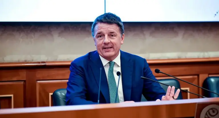 Un uomo in abito parla durante una conferenza stampa, seduto a un tavolo con microfoni